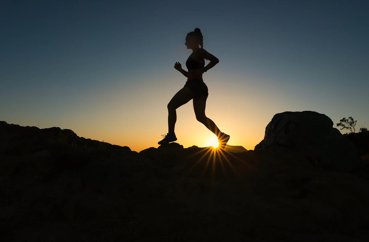 A woman in silhouette running on a trail against a low sun